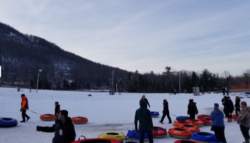 People walking on a snowy hill with colorful snow tubes scattered around, surrounded by trees and mountains.