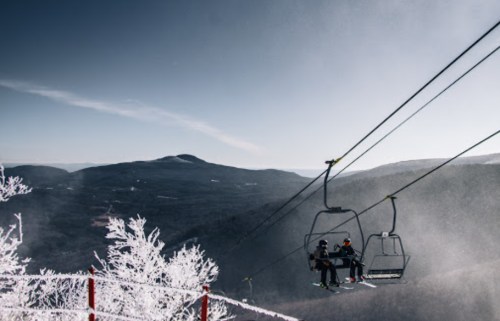 Two skiers on a chairlift, surrounded by snowy mountains and a clear blue sky.