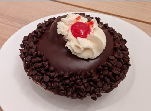 A round chocolate cake topped with whipped cream and a cherry, surrounded by chocolate chips on a white plate.