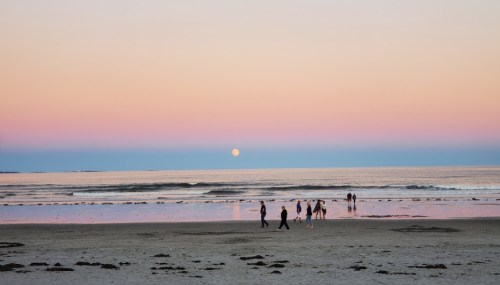 Silhouettes of people walking on a beach at sunset, with a full moon rising over calm waters and a pastel sky.