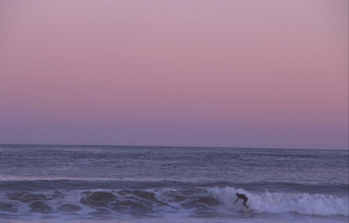 A surfer rides a wave at sunset, with a pastel pink sky and calm ocean in the background.