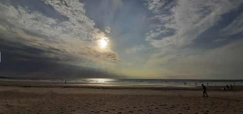 A serene beach scene at sunset, with gentle waves, a cloudy sky, and a few people walking along the shore.