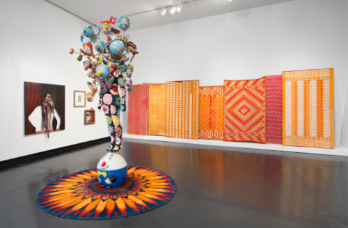 Colorful art installation featuring a tree-like sculpture and vibrant patterned wall hangings in a gallery setting.