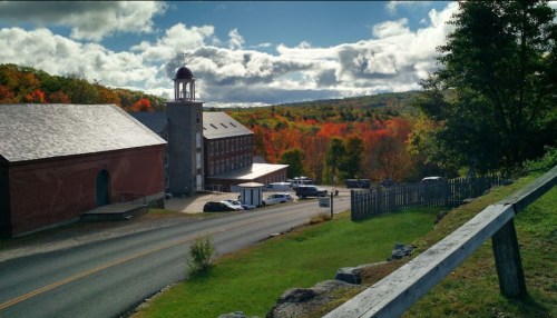 A scenic view of a red building beside a road, with vibrant autumn foliage in the background and a cloudy sky.