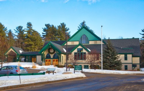 A modern church building with green accents, surrounded by snow and trees, under a clear blue sky.