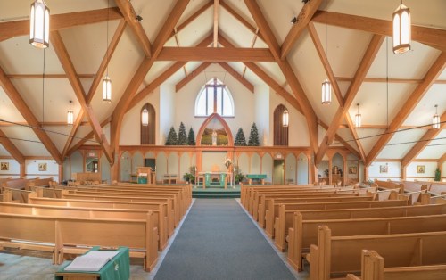 Interior of a church featuring wooden beams, pews, and an altar, with natural light streaming through large windows.