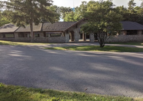 A stone building with a sloped roof surrounded by trees, viewed from a road on a sunny day.