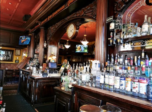 A wooden bar with a variety of liquor bottles displayed, a clock on the wall, and TVs showing sports in a cozy pub setting.