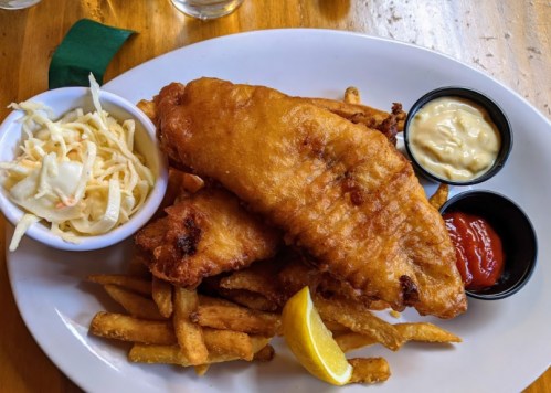 A plate of crispy fried fish fillets with fries, coleslaw, lemon wedge, and dipping sauces.