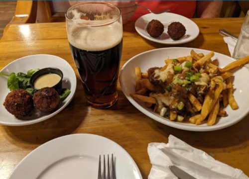 A wooden table with a pint of dark beer, loaded fries topped with cheese and green onions, and two plates of appetizers.