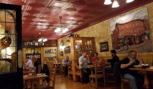 A cozy restaurant interior with wooden tables, patrons dining, and a mural on the wall depicting a street scene.