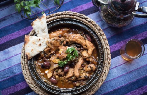A traditional dish of chicken with olives, served in a clay pot, accompanied by flatbread and a glass of tea.