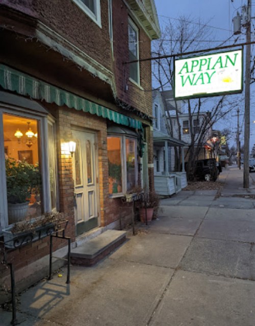 A cozy brick building with a green awning and a sign reading "APPIAN WAY," illuminated in the evening.