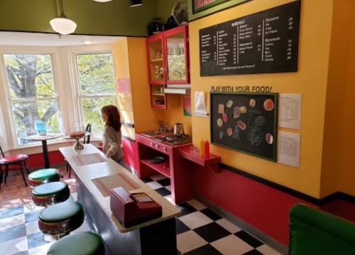 A colorful diner interior with a counter, red cabinets, a chalkboard menu, and a woman standing by the stove.