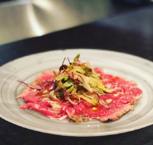 A plate of thinly sliced beef carpaccio topped with a mix of greens and drizzled with dressing.