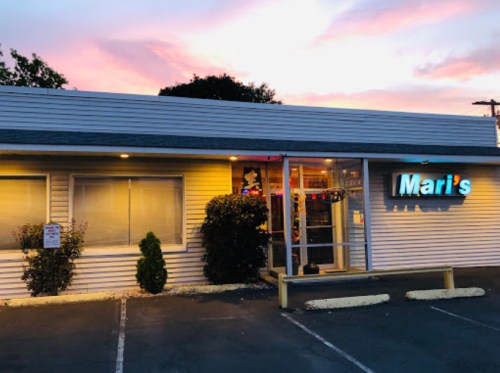 A small restaurant named "Mari's" with a colorful sign, surrounded by greenery and a sunset sky in the background.