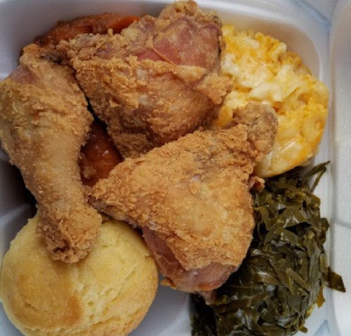 A takeout container with fried chicken, macaroni and cheese, cornbread, collard greens, and a side of sauce.