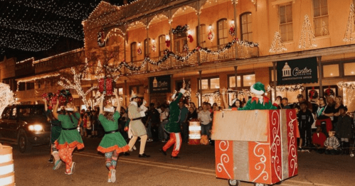 A festive parade scene with performers in holiday costumes, decorated buildings, and a crowd enjoying the celebration.