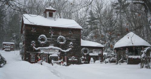 A snowy scene of a rustic building decorated with wreaths and holiday ornaments, surrounded by trees.