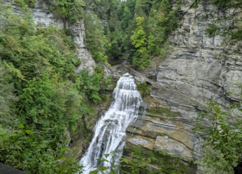 A cascading waterfall flows through a rocky gorge, surrounded by lush green trees and steep cliffs.
