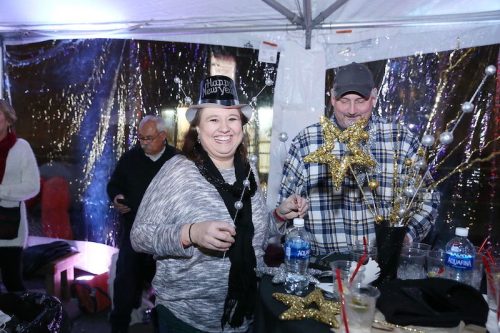 A woman in a "Happy New Year" hat smiles at a festive gathering, while a man in a plaid shirt stands nearby.