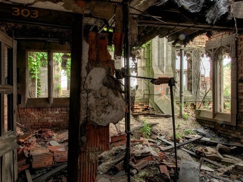 A dilapidated interior of a ruined building, with exposed brick, debris, and overgrown vegetation visible through broken windows.