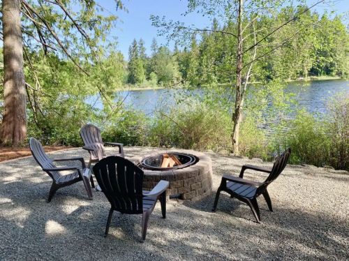 A serene lakeside scene featuring a fire pit surrounded by four chairs, framed by trees and greenery.