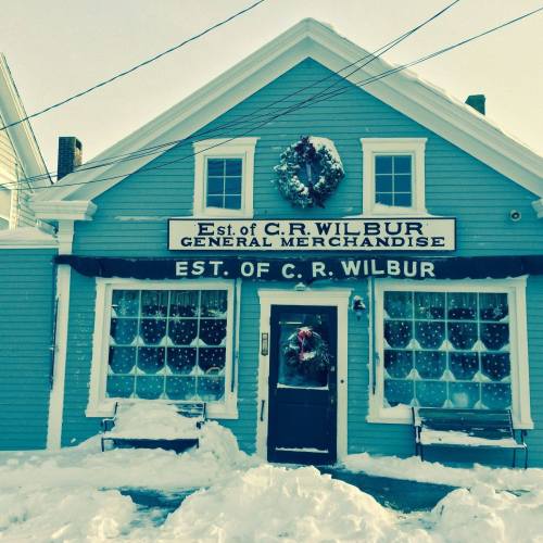 A blue building with a sign reading "Est. of C.R. Wilbur General Merchandise," decorated for winter and covered in snow.