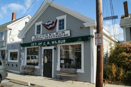 A quaint general merchandise store with a sign reading "C.R. Wilbur" and patriotic decorations on a sunny day.