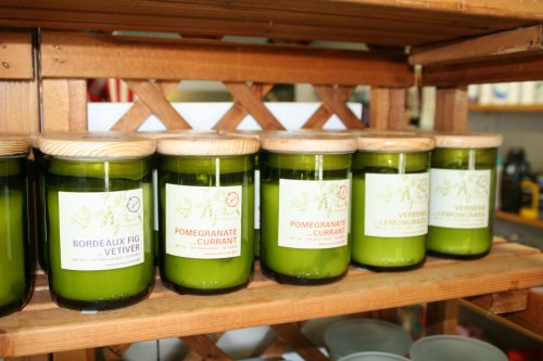 A wooden shelf displaying several green candle jars with labels for Bordeaux Fig Vetiver, Pomegranate Currant, and more.
