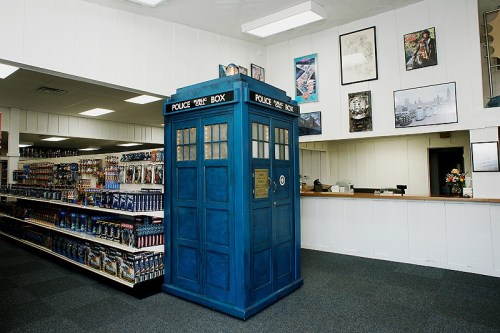A blue police box stands in a store, surrounded by shelves filled with various merchandise and posters on the walls.