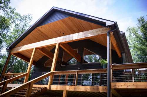 A modern cabin with a wooden deck and large windows, surrounded by trees under a clear blue sky.