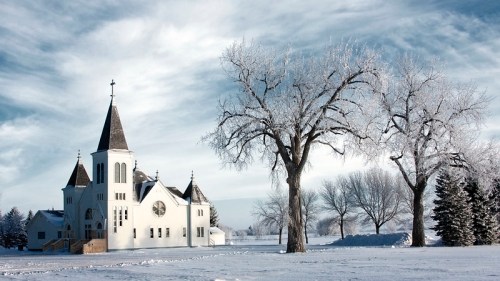 A white church with a tall steeple surrounded by snow-covered trees and a winter landscape.