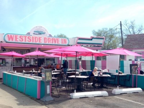 A colorful drive-in diner with pink umbrellas and outdoor seating, featuring a retro design and a vibrant atmosphere.