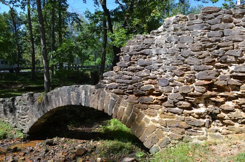 A weathered stone bridge arching over a small stream, surrounded by trees and greenery.