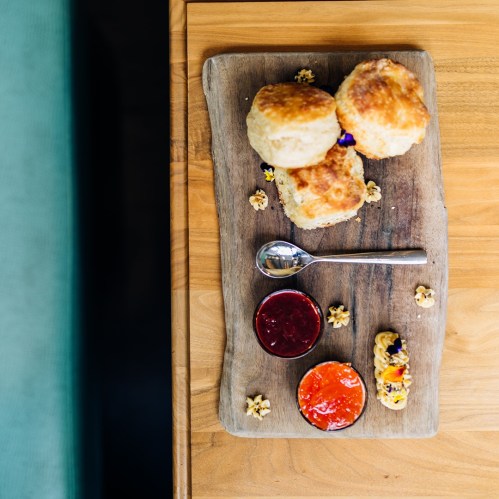 A wooden platter with three scones, two jams, and a decorative cookie, surrounded by small edible flowers.