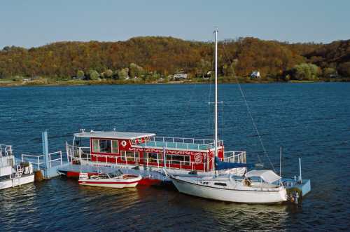 A red and white houseboat docked alongside a sailboat on a calm river, with autumn trees in the background.