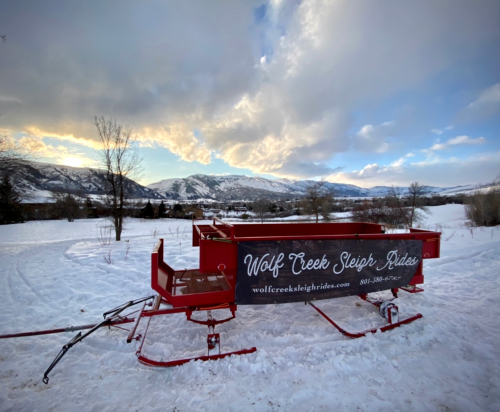 A red sleigh with "Wolf Creek Sleigh Rides" on it, set against a snowy landscape and mountains under a cloudy sky.