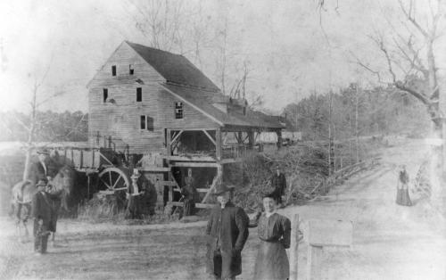 Black and white photo of a historic mill with people and horses nearby, set in a rural landscape.