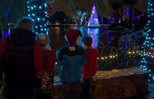 Children in winter attire admire a brightly lit Christmas tree by a lake, surrounded by festive decorations.