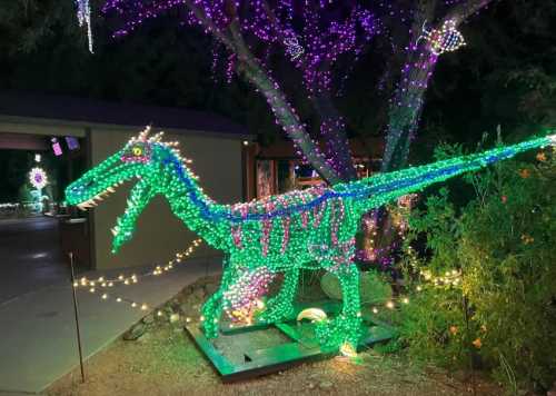 A brightly lit dinosaur sculpture made of green and purple lights, surrounded by festive decorations in a garden setting.