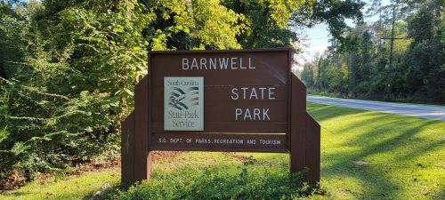 Sign for Barnwell State Park in South Carolina, surrounded by trees and greenery, with a road visible in the background.