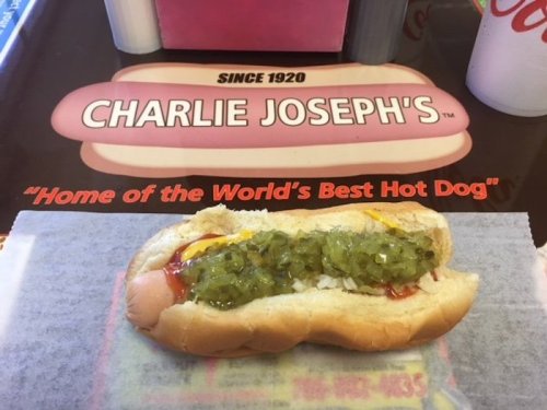 A hot dog topped with mustard, ketchup, and relish on a paper wrapper, with a sign for Charlie Joseph's in the background.