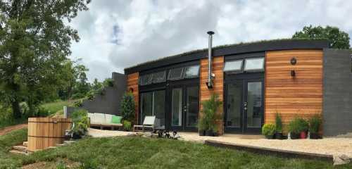 A modern home with a green roof, wooden exterior, large windows, and a patio surrounded by greenery.