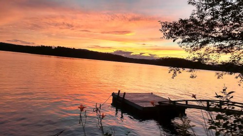 A serene lake at sunset, with vibrant orange and pink skies reflecting on the water and a small dock in the foreground.