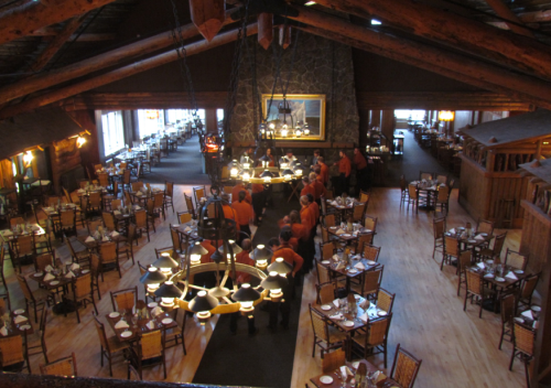 A spacious restaurant with wooden beams, tables set for dining, and staff gathered around a large central table.