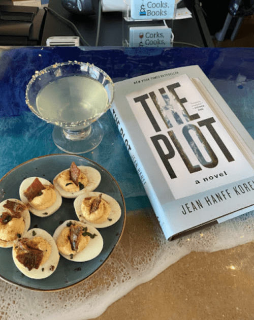 A cocktail, a plate of appetizers, and a novel titled "The Plot" on a colorful table.