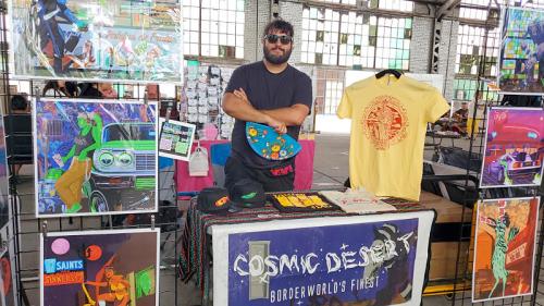 A vendor stands behind a table displaying colorful artwork and T-shirts at a market.