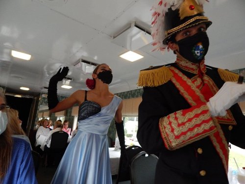 A performer in a blue gown and a masked man in a military-style uniform dance in a train car filled with people.