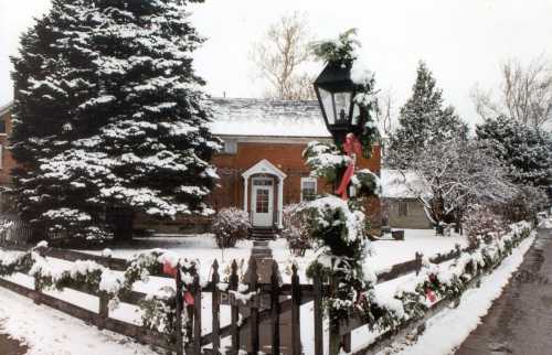 A snowy scene of a brick house with a lamppost, surrounded by a festive fence adorned with greenery and ribbons.